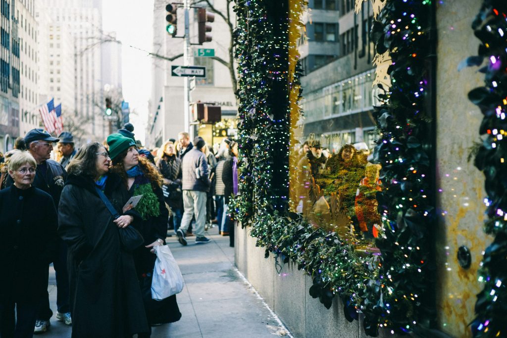 A line of people stands on a city sidewalk, admiring a vibrant window display filled with various items.
