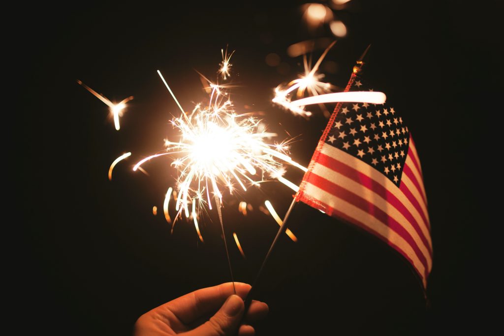 A hand holds a sparkler, illuminating the night, with an American flag waving in the background, symbolizing patriotism.