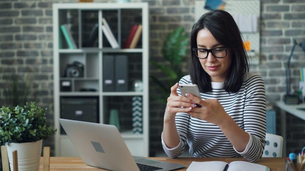A woman in glasses sitting at a desk with a laptop and cell phone